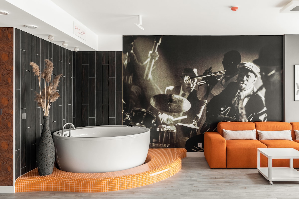 A spacious bathroom area features a round, freestanding jacuzzi bathtub on an elevated platform. A modern orange sofa is visible nearby, accompanied by a minimalist white coffee table. A large black and white mural depicting musicians creates an artistic backdrop, enhancing the contemporary design.