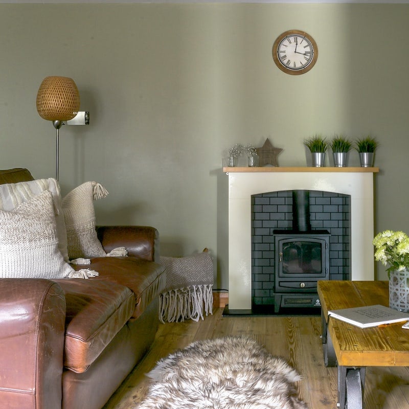 A cozy lounge area is captured, featuring a leather sofa adorned with textured cushions. A simple wooden coffee table sits in front, with a decorative vase holding flowers. The black stove adds warmth, framed by a minimalist mantle featuring small potted plants and a wall clock.