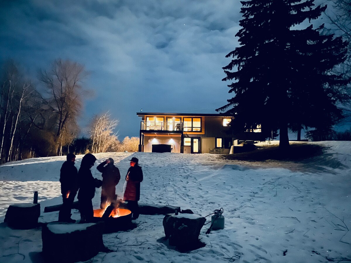 A cozy outdoor gathering is taking place around a fire pit in a snowy landscape, with four individuals enjoying the warmth of the flames. The remodeled home is visible in the background, illuminated softly against the evening sky.