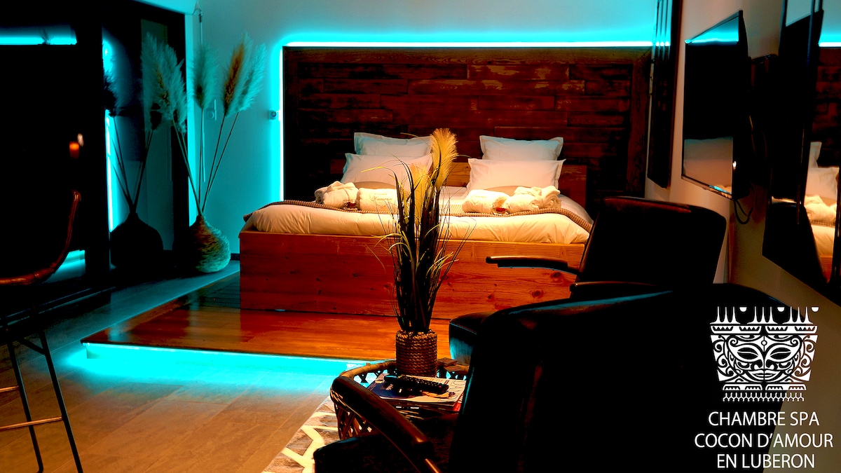 A modern bedroom features a spacious bed with a wooden headboard, illuminated by soft blue lighting. Decorative plants are positioned in the room, while a cozy seating area provides additional comfort. The overall atmosphere is serene and designed for relaxation.