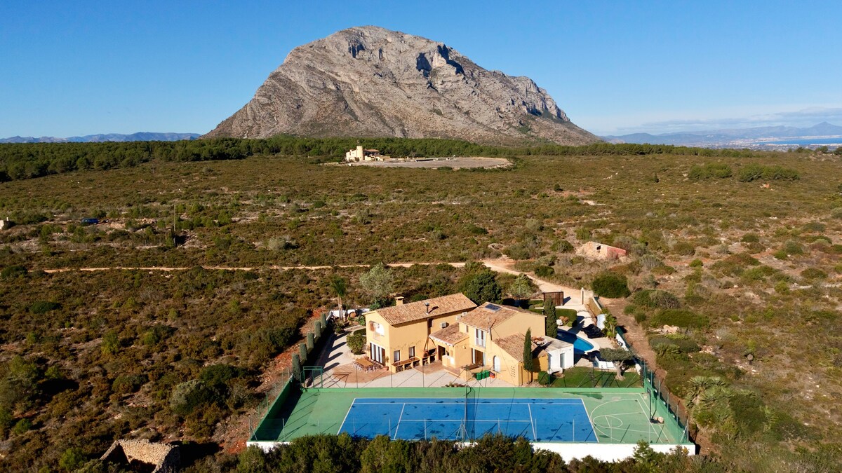 A spacious villa is situated amidst natural landscapes, with a large tennis court in front. The Montgó mountain rises majestically in the background under a clear blue sky, emphasizing the tranquility of the surrounding area.