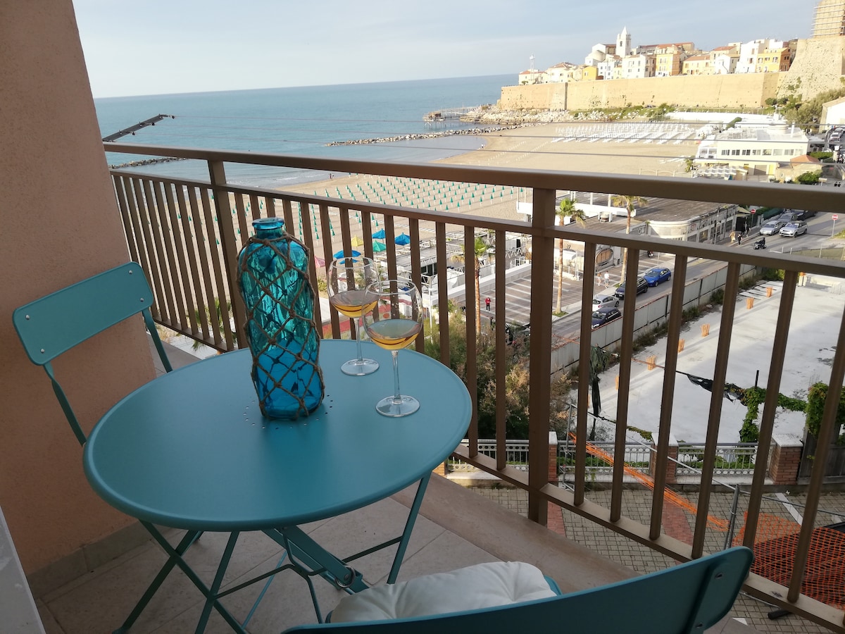 A small terrace features a round blue table and two matching chairs, overlooking the sea. A decorative blue glass vase is placed on the table, with two glasses of wine positioned nearby. The view includes the beach and historic buildings in the background.