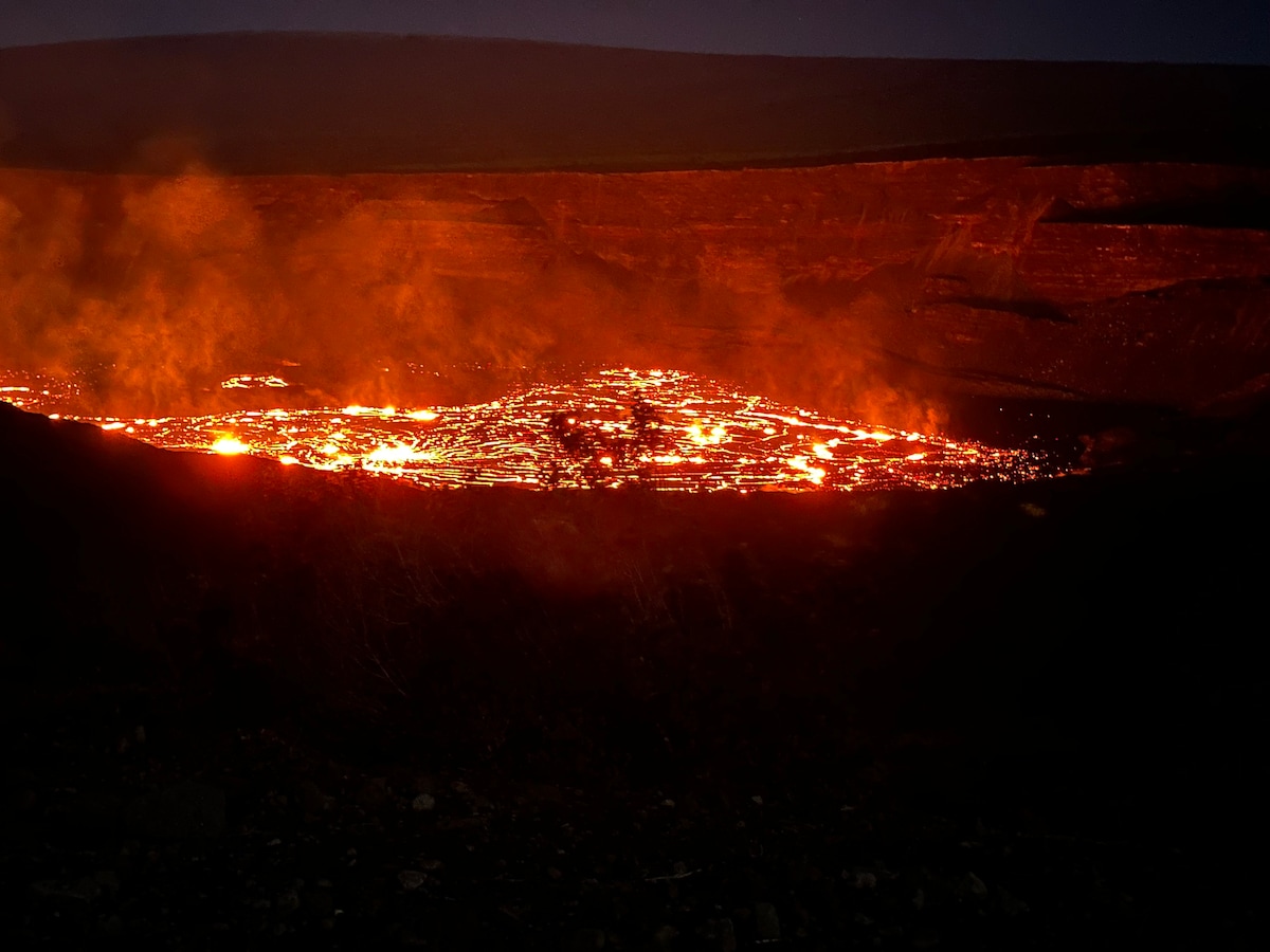 A glowing view of lava flows is showcased against a dark landscape, illuminating the surrounding area with warm orange and red hues. Smoke rises gently from the surface, creating a captivating contrast with the black volcanic terrain.