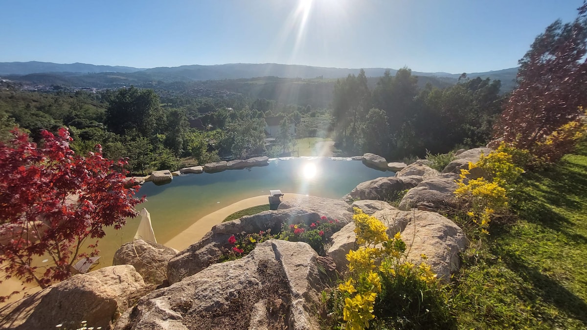 A serene pool area is framed by natural stone and vibrant foliage, offering views of the surrounding hills. Sunlight reflects off the water's surface, creating a tranquil atmosphere. Rich greenery and blooming flowers enhance the outdoor space, contributing to a peaceful retreat.