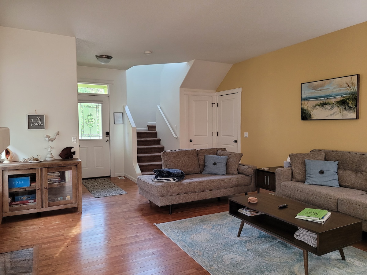 The living room features two light brown sofas facing each other, with a wooden coffee table positioned between them. A staircase leads to the upper floor, and light enters through the front door. Neutral walls are complemented by a small piece of artwork.