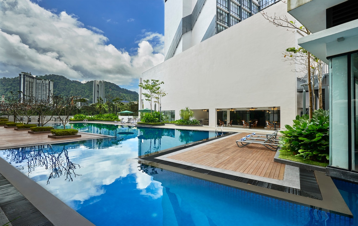 An expansive swimming pool features clear blue water, reflecting the surrounding palm trees and mountains. Lounge chairs are arranged alongside the deck, which is made of light wood. The calm environment invites relaxation, with green landscaping framing the pool area and modern architecture in the background.