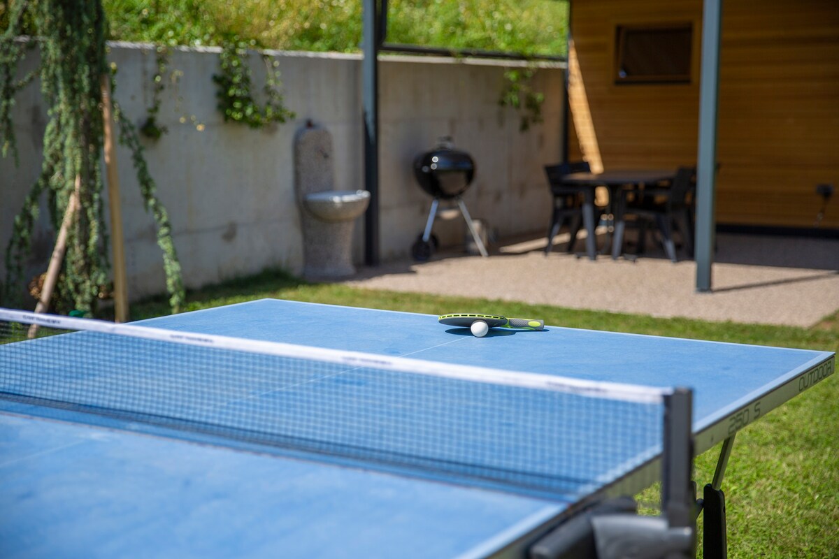 A blue table tennis table occupies a section of the large garden, with a paddle and a ball resting on it. In the background, a Weber grill is visible next to a covered dining area, providing a space for outdoor meals.