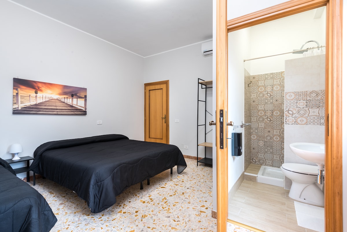 A spacious bedroom features two dark beds with black bedding. A large window allows natural light to fill the room. A modern bathroom is visible in the background, showcasing patterned tiles and a clear shower enclosure. A simple bedside table stands beside one bed.
