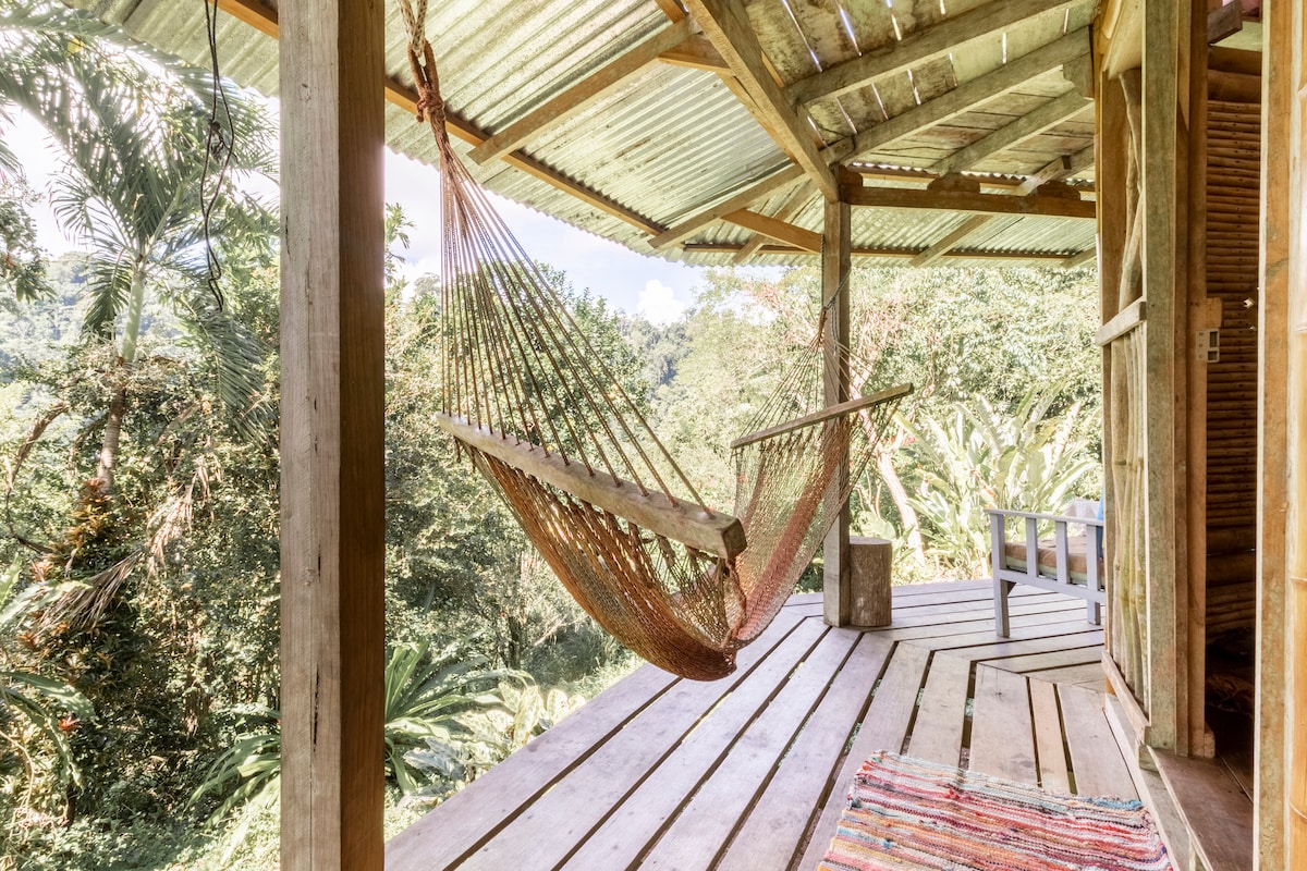 A spacious wooden deck features a relaxing hammock suspended from the ceiling, surrounded by lush greenery. The natural wood flooring complements the serene setting, while a soft, colorful textile adds a touch of warmth. Expansive views of the surrounding forest create a tranquil atmosphere.