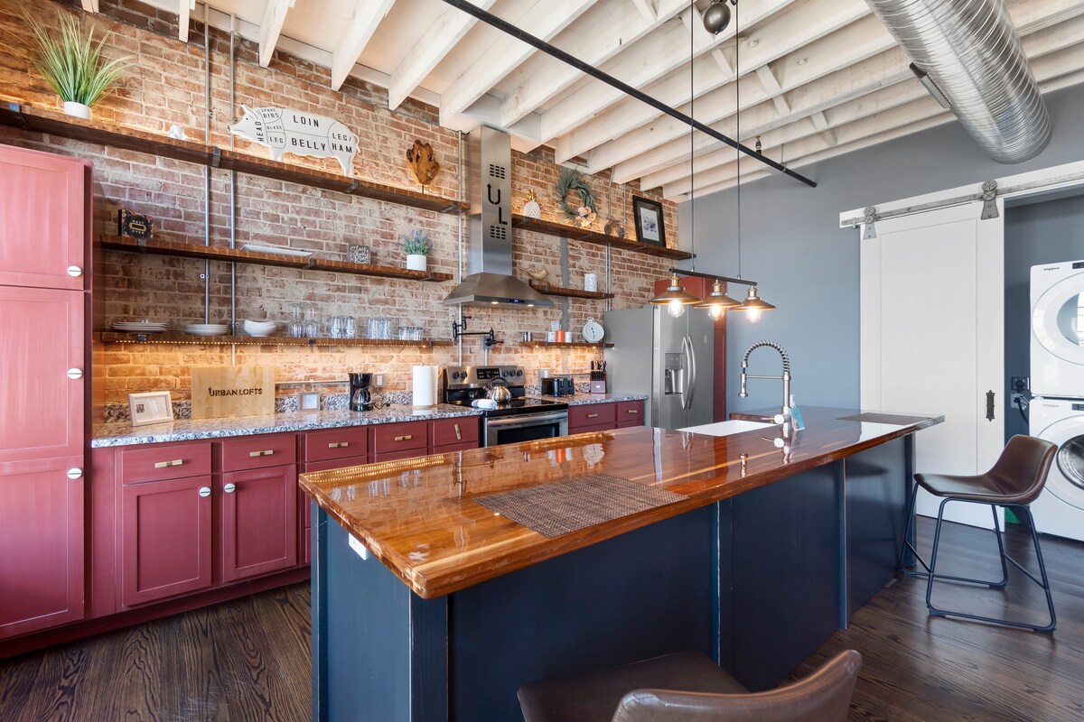 A modern kitchen with exposed brick walls features dark wooden cabinetry and a large island with a polished wooden countertop. Stainless steel appliances are visible, along with open shelving displaying decorative items. Natural light highlights the space, enhancing the inviting atmosphere.