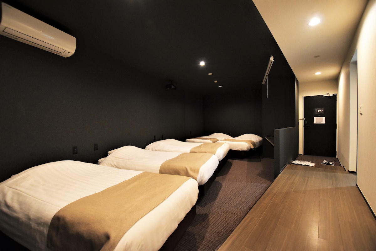 A private room is equipped with several spacious beds, arranged in a minimalist layout. The room features a dark, modern color scheme complemented by warm lighting. A door leads to the outside corridor while a mirror reflects part of the tranquil space.