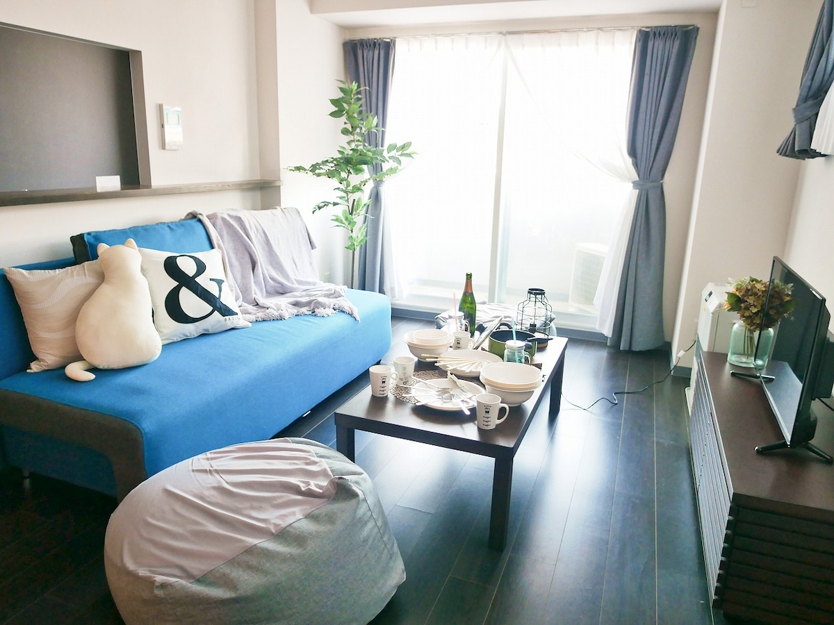 The living area is adorned with a blue sofa complemented by neutral decorative pillows. A wooden coffee table sits in front, adorned with plates and cups. Natural light filters through the sheer curtains, illuminating the space. A small plant adds a touch of greenery to the room.