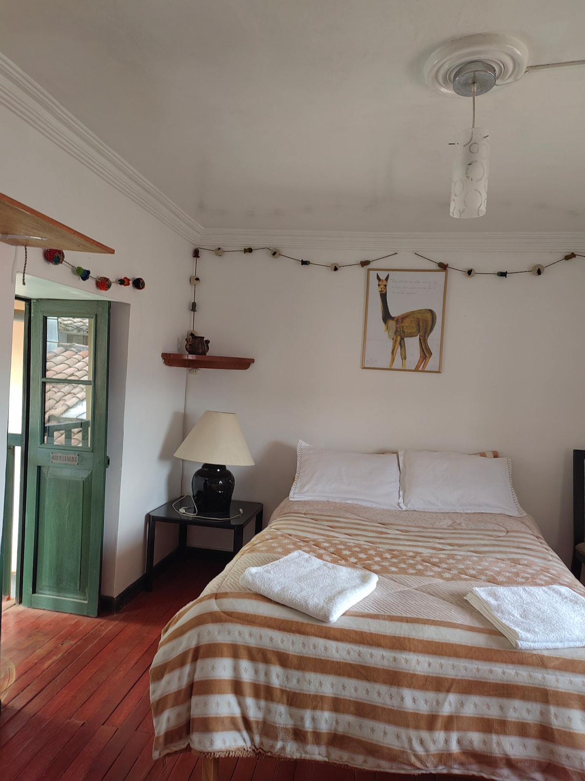 A cozy bedroom features a spacious bed with cream-colored linens. Natural light fills the room through a green door, leading to the outside. Decorative string lights hang along the wall, and a framed illustration of a llama is displayed above a nightstand with a lamp.