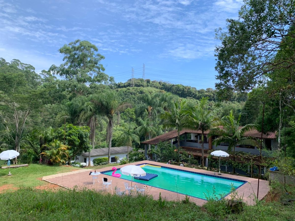 A spacious outdoor area features a large swimming pool, surrounded by lush greenery and palm trees. White umbrellas provide shade over several loungers. In the background, a two-story house complements the natural landscape, with its warm tones blending with the surroundings.