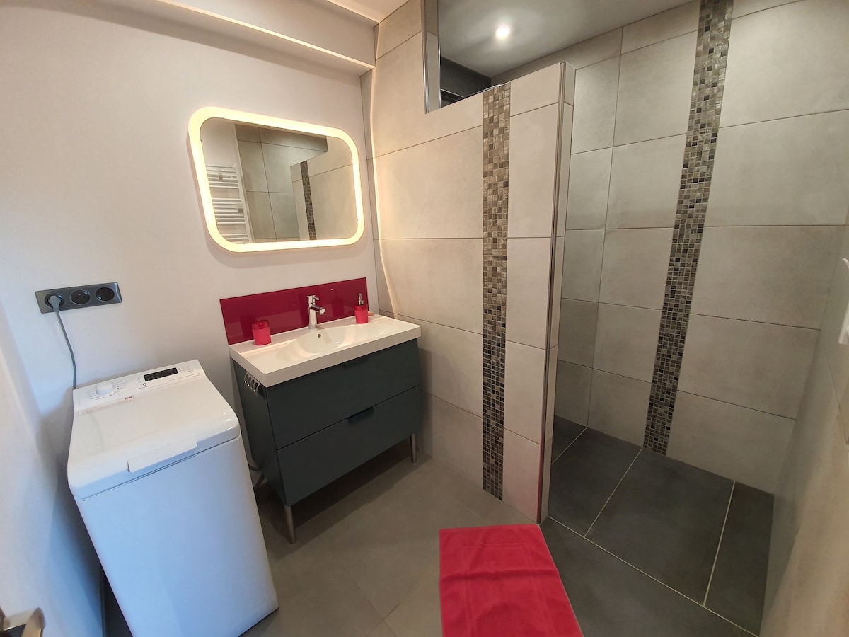 A modern bathroom features a spacious walk-in shower with tiled accents and a sleek vanity with dual sinks. A compact washing machine is situated nearby. Soft lighting is integrated into the design, alongside a large mirror, creating an open and functional space.