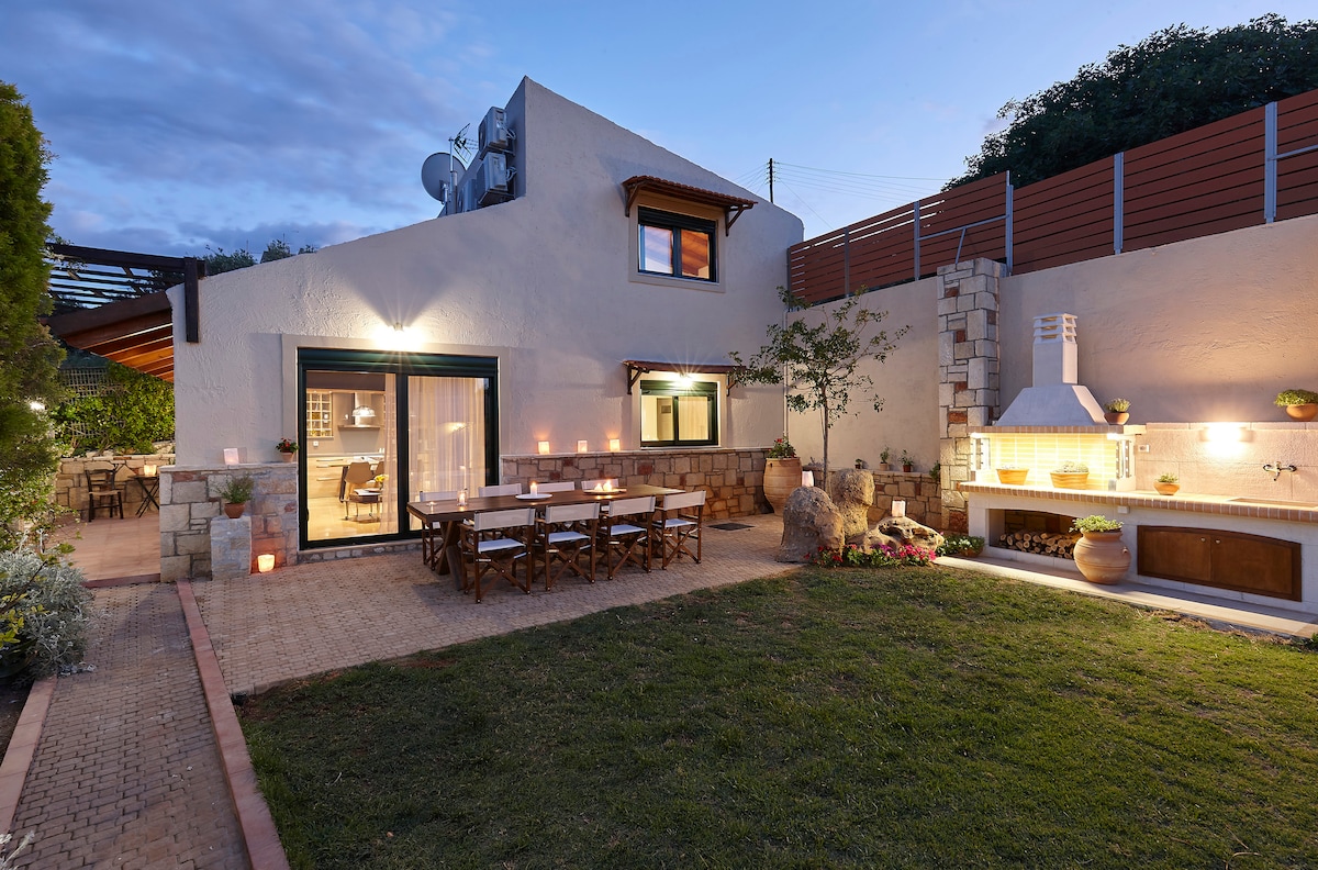 The exterior of Villa Thallos is illuminated by soft outdoor lighting. A spacious patio area features a wooden dining table surrounded by chairs, with a brick barbecue setup nearby. Lush greenery and garden elements enhance this inviting outdoor space.