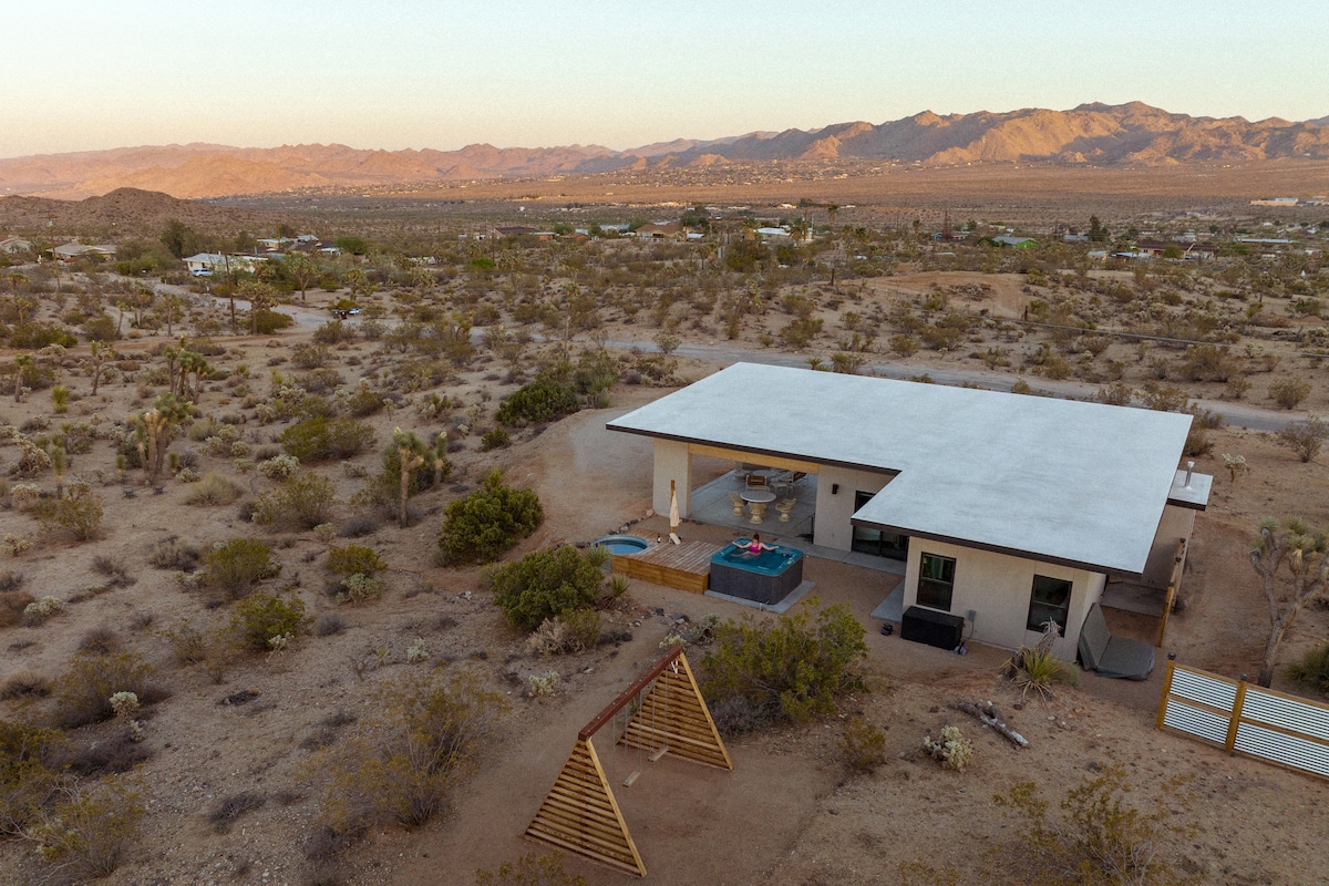 An aerial view of Mojave Rosa reveals a mid-century modern home surrounded by desert landscape. A blue hot tub and lounge area are visible along with a custom swing set in the outdoor space, providing a relaxing atmosphere with mountains in the background.