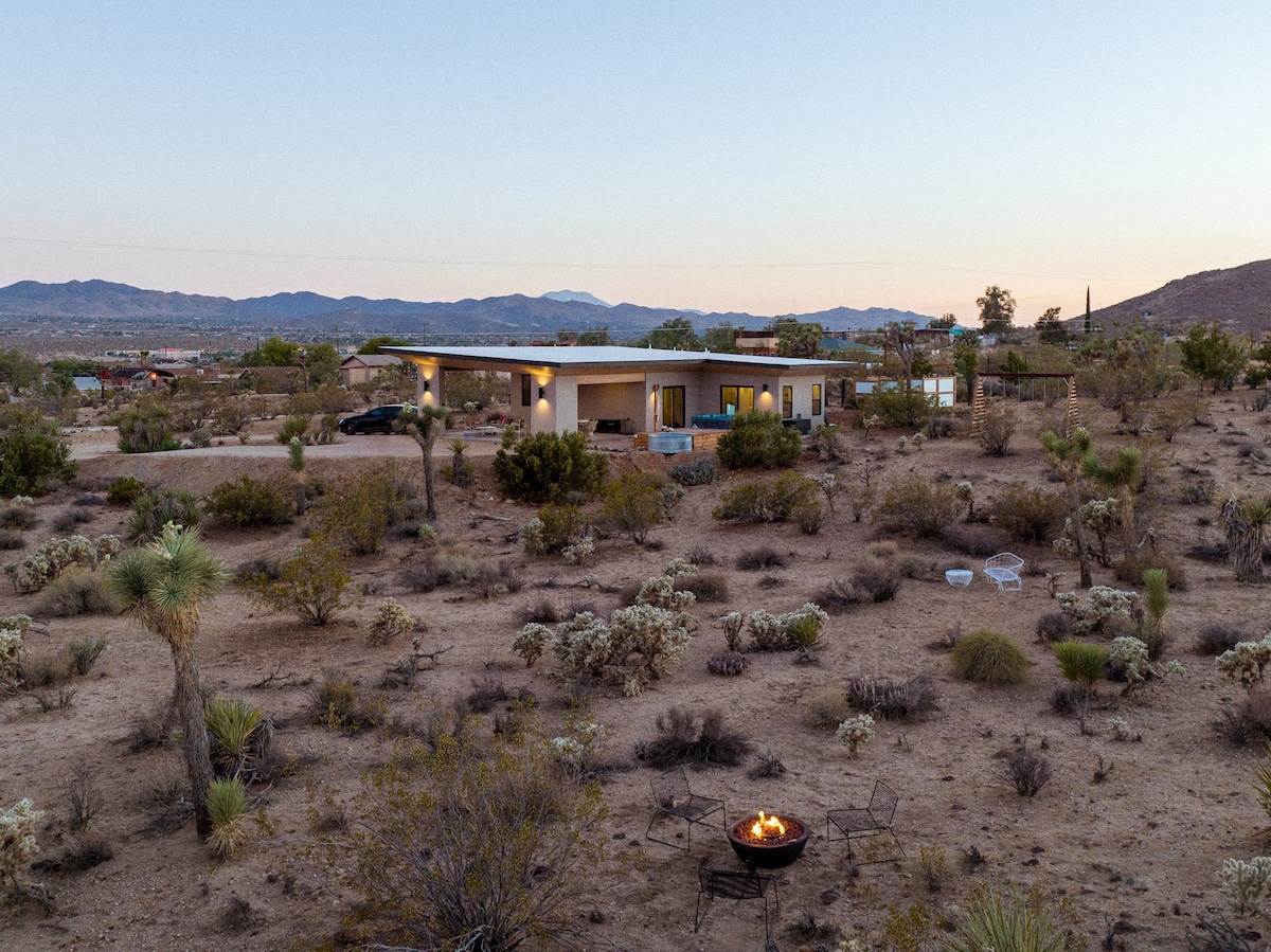 The home is surrounded by desert vegetation, presenting a blend of rugged terrain and modern architecture. A firepit is situated in the foreground, inviting for evening gatherings. The distant mountains fade into the twilight, enhancing the serene ambiance of the landscape.