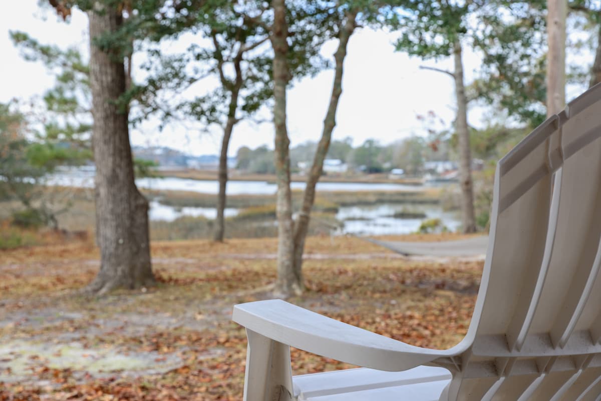 A white Adirondack chair is positioned in the foreground, offering a view of tranquil waters framed by trees. The natural landscape features soft earth tones with fallen leaves scattered across the ground, creating a peaceful setting near the river.