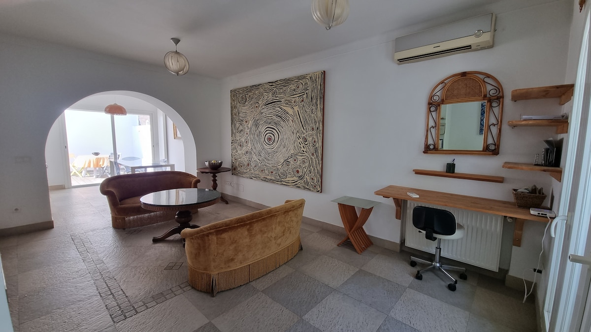 A modern living area features curved sofas around a circular coffee table, with an artistic wall hanging adding character. A workstation with a chair is present, along with a large mirror on the wall. Natural light brightens the space through adjoining openings.