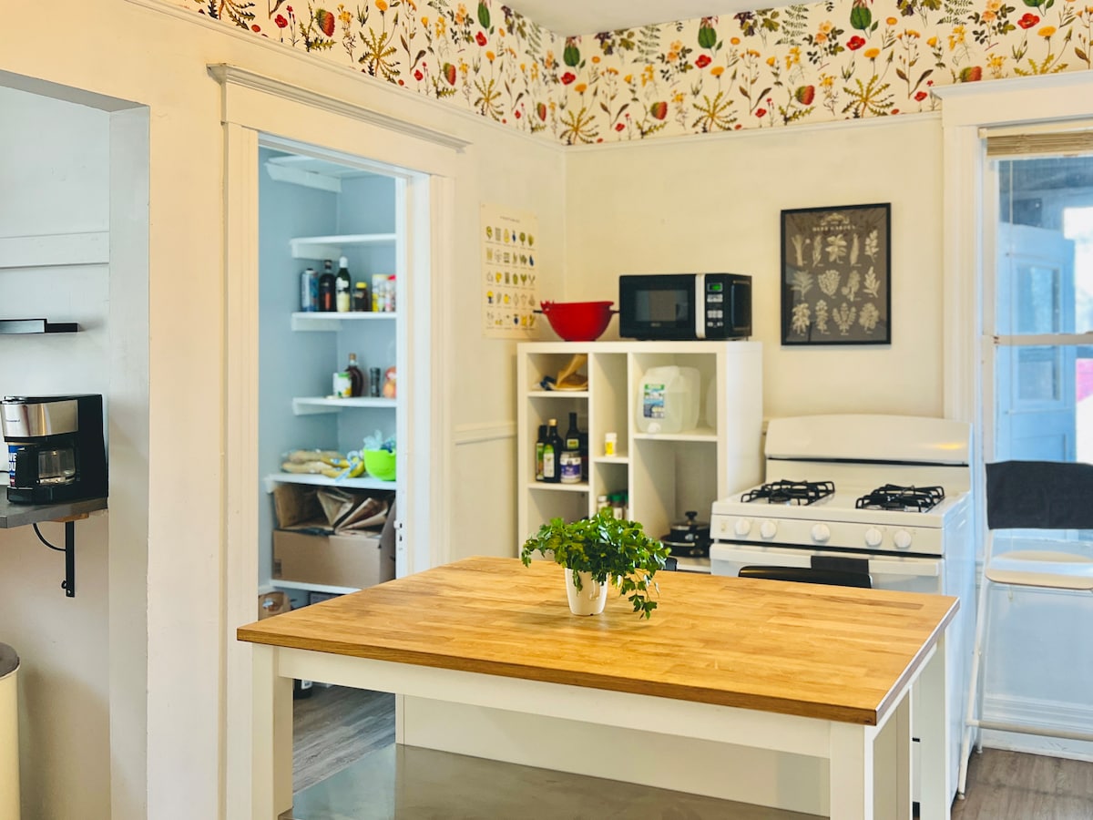 A bright kitchen features a central wooden island with a potted plant on top. White cabinetry lines the walls, and a gas stove is positioned next to a microwave and open shelving stocked with pantry items. Floral wallpaper adds a cheerful touch.