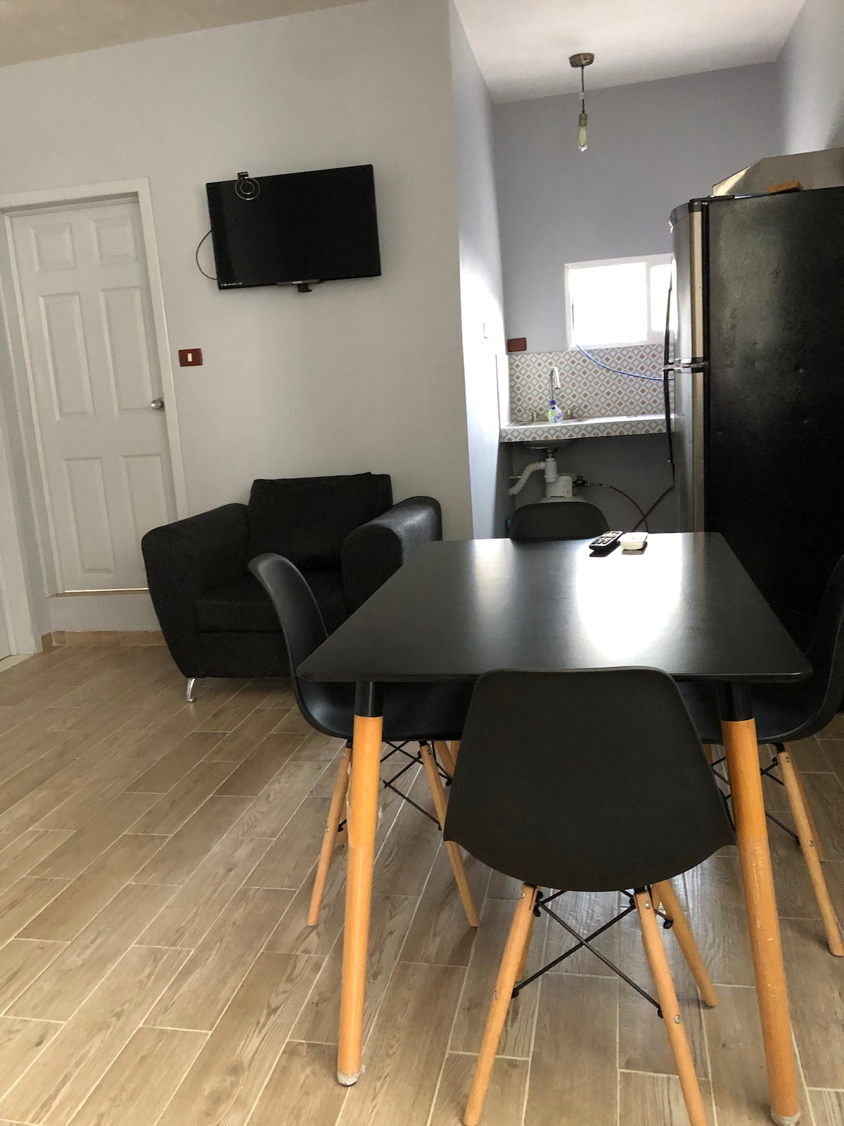 A cozy dining area features a black table surrounded by four chairs, complemented by a small black sofa in the corner. A television is mounted on the wall, while the kitchen area is visible in the background, showcasing a refrigerator and sink.