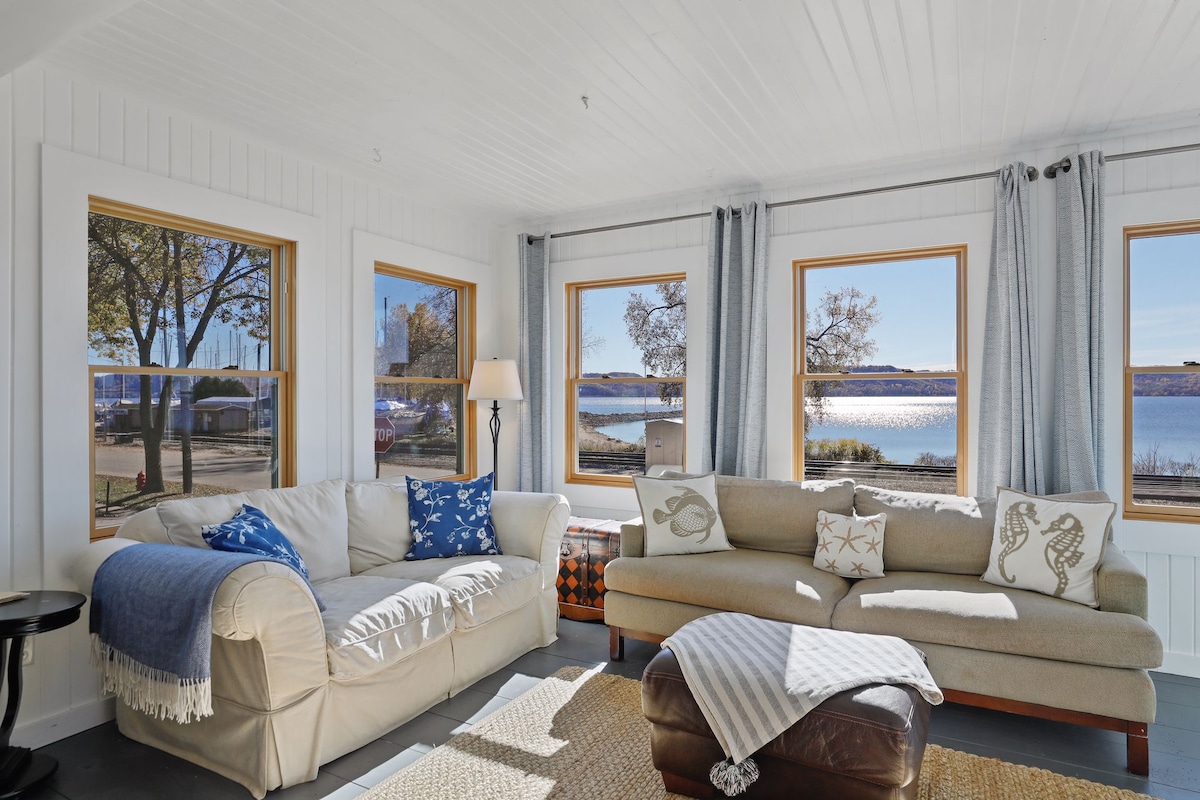 A bright sunroom features large windows offering views of the lake. A light-colored couch is adorned with decorative pillows, and a cozy armchair is positioned nearby. Natural light fills the space, highlighting the warm tones of the furnishings and the textured area rug beneath.