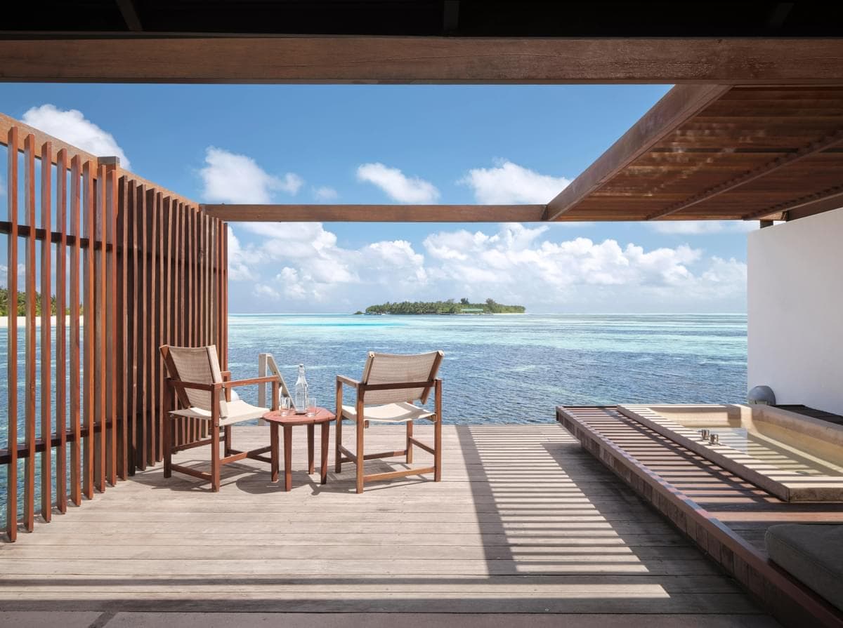 A private terrace features two cushioned chairs and a small table, overlooking the lagoon. The tranquil water reflects a clear blue sky with soft clouds, while a distant island is visible, enhancing the serene atmosphere of the space.