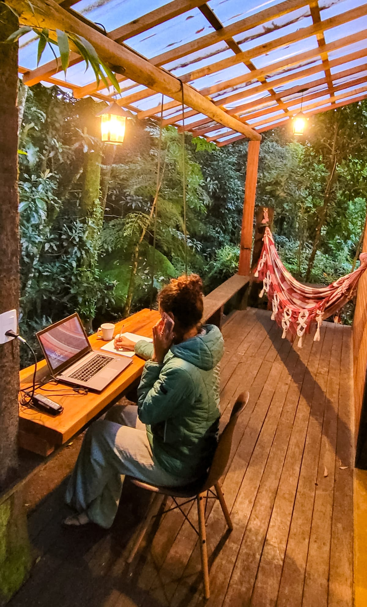 A wooden deck is shown, featuring a seated individual working on a laptop, surrounded by lush greenery. Soft lighting from hanging lamps creates an inviting atmosphere. A hammock is visible in the background, offering a cozy spot for relaxation.