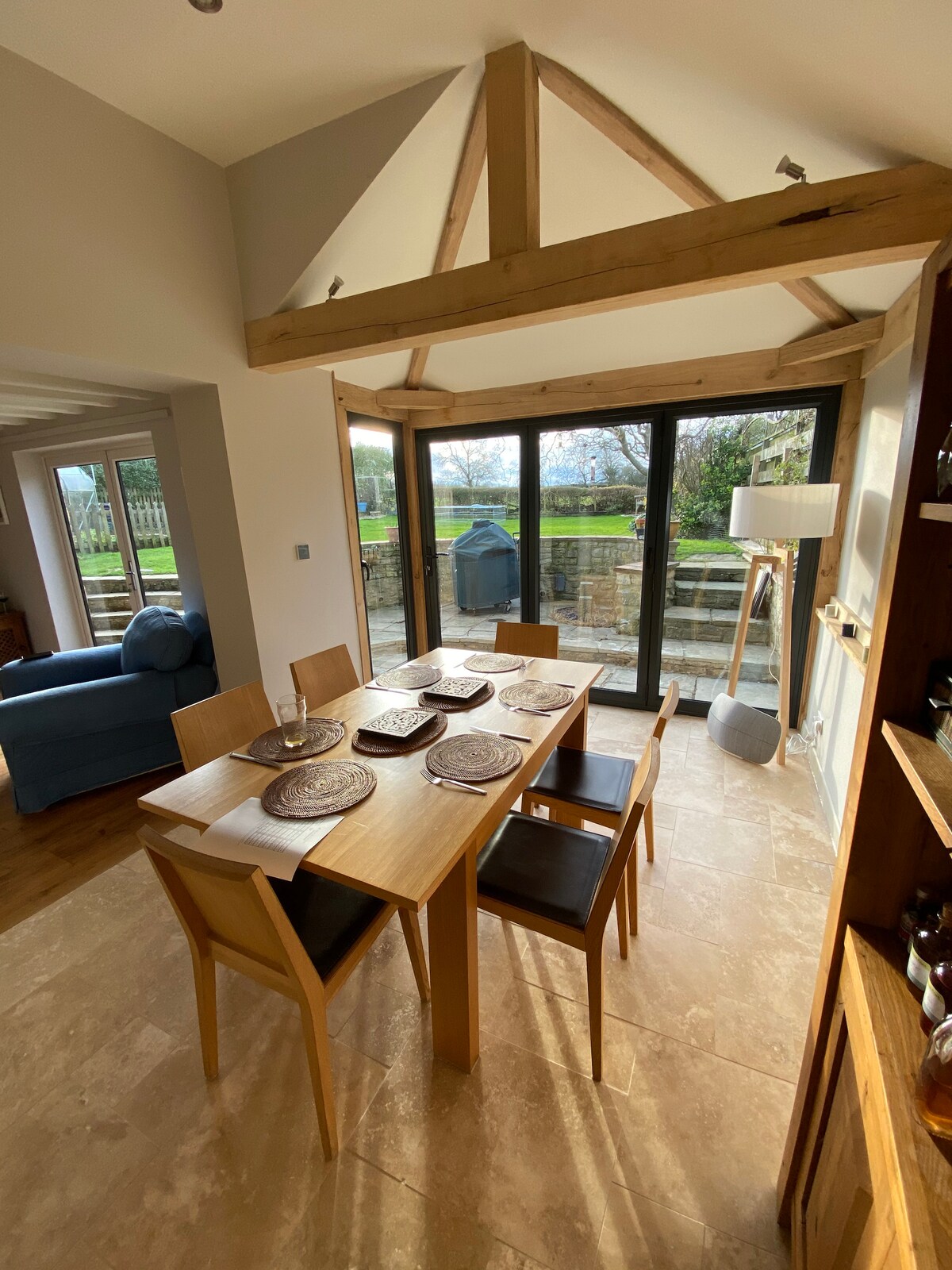 A dining area features a wooden table set for four with woven placemats. Large glass doors provide views of the green landscape outside, enhancing natural light. Exposed wooden beams line the ceiling, contributing to an airy atmosphere. A lamp and a cozy seating arrangement are also visible.