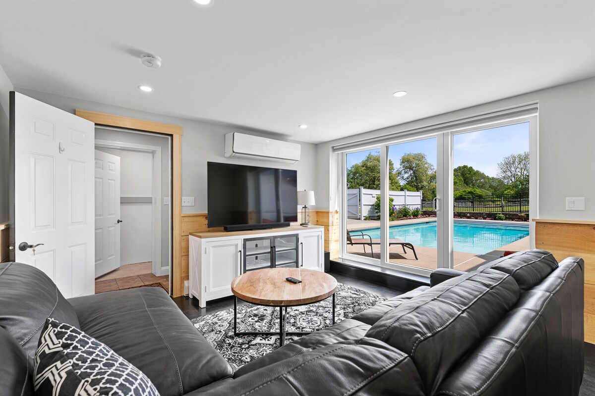 A spacious living area is equipped with a large television and a comfortable sofa. Natural light floods in through expansive windows that offer views of the outdoor pool area. A round coffee table stands in the center, enhancing the room's functionality.