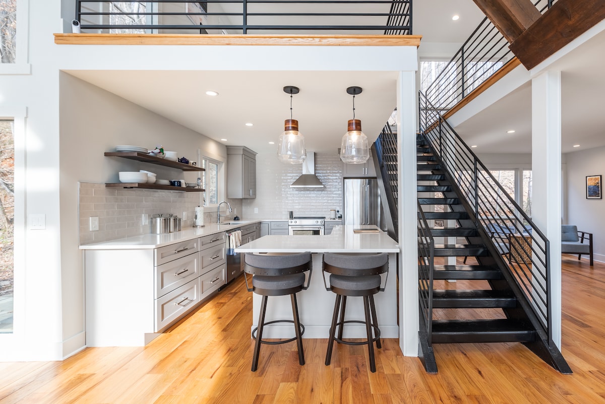 A modern kitchen features sleek cabinetry, a spacious island with two stools, and pendant lighting. Large windows allow natural light to illuminate the bright interior. The adjacent open staircase leads to the loft area, showcasing the home's contemporary design.