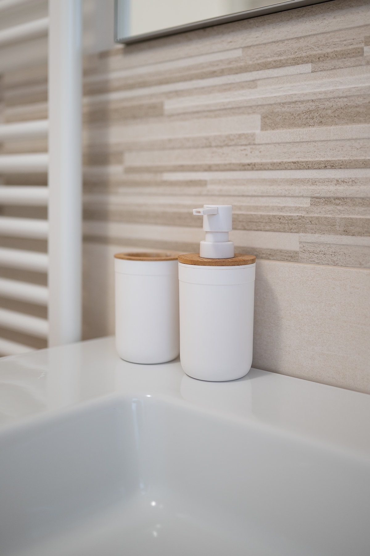 A sleek bathroom countertop is shown, featuring two white containers with wooden lids. One container is equipped with a pump dispenser. The backdrop consists of textured beige and light grey tiling, contributing to a clean and modern aesthetic.