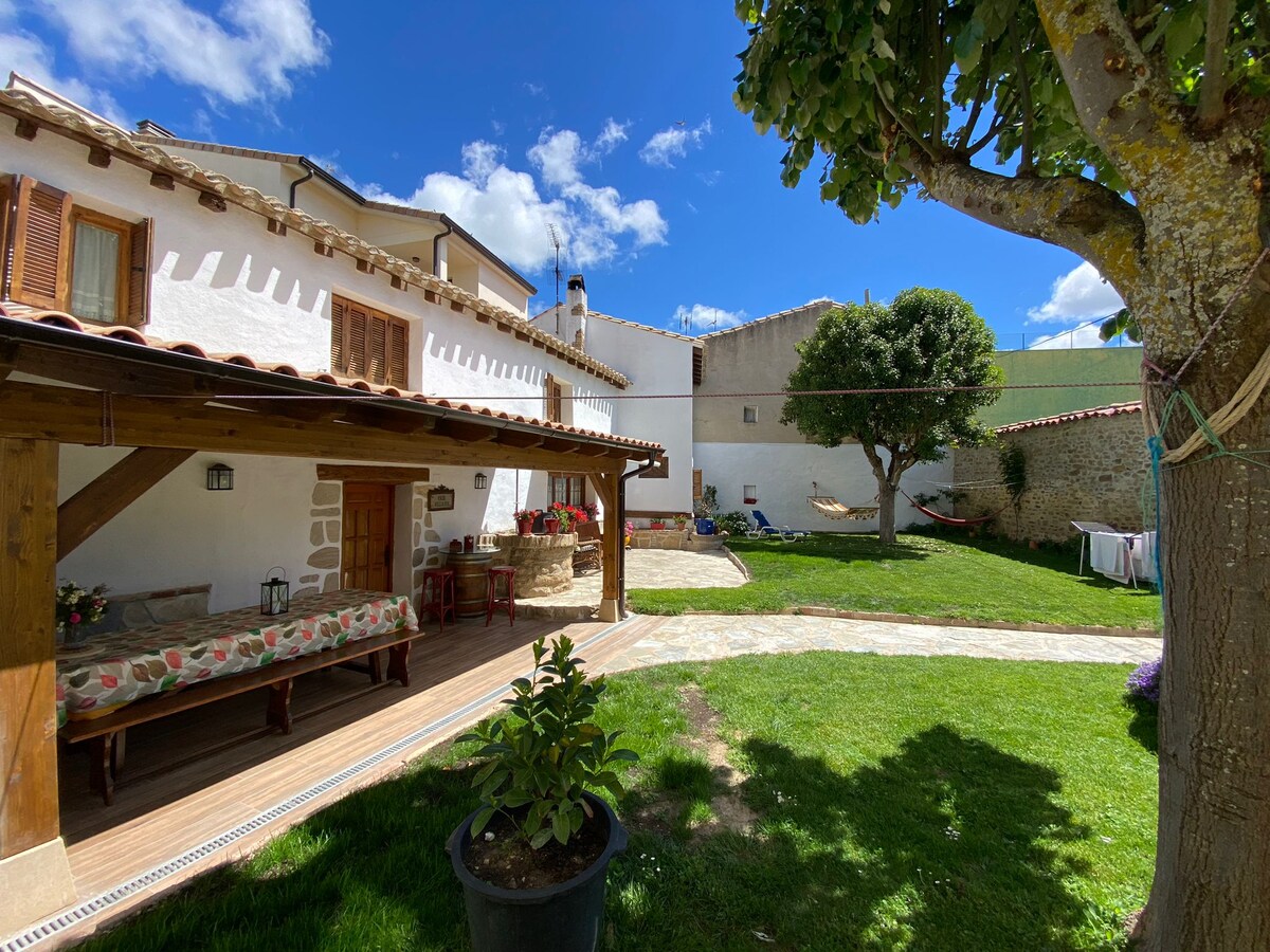 A spacious outdoor area features a wooden terrace with a floral-patterned seating arrangement. Lush green grass is surrounded by a stone path and a tree, providing shade. A hammock is visible in the background, and laundry hangs nearby, indicating a welcoming residential setting.