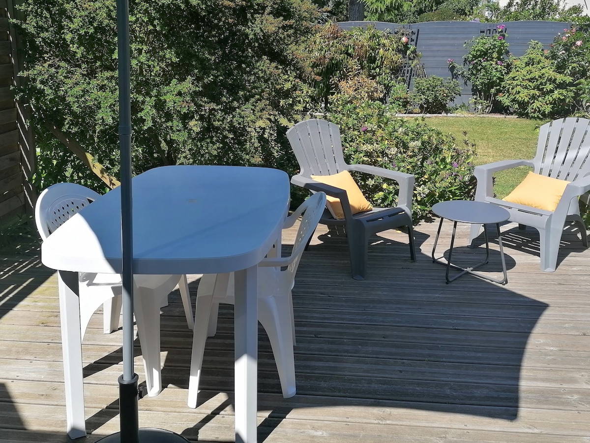 A private terrace is presented, featuring a white table surrounded by four chairs and two lounge chairs with cushions. The space is shaded by a parasol and is bordered by greenery, creating a serene outdoor setting.