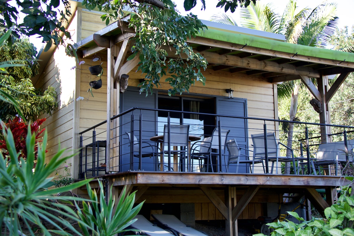 A wooden bungalow is surrounded by tropical greenery, featuring a spacious terrace with a dining table and several chairs. Large windows provide access to natural light and views of the surrounding garden. The overhanging roof offers shaded seating areas.