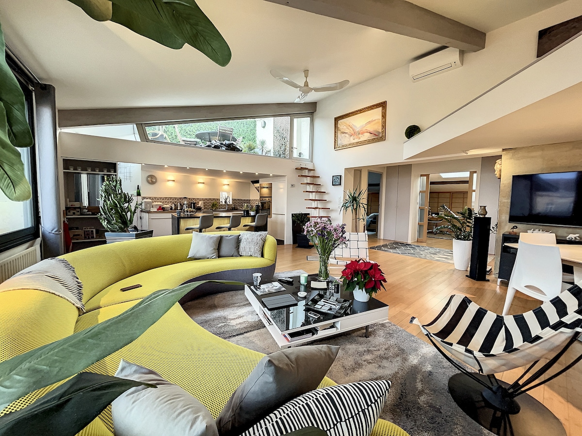 The spacious living area features a bright yellow curved sofa arranged around a low coffee table. A large television is positioned on a wall, while a staircase leads to an upper level. Generous natural light filters through large windows, complemented by indoor plants and decorative items.