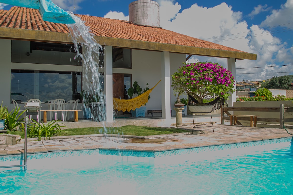 A tranquil outdoor area features a swimming pool with a waterfall cascading into it. Colorful flowers frame the pool, while two hammocks are positioned nearby. The house's large glass doors reveal a spacious living area, enhancing the connection to nature.