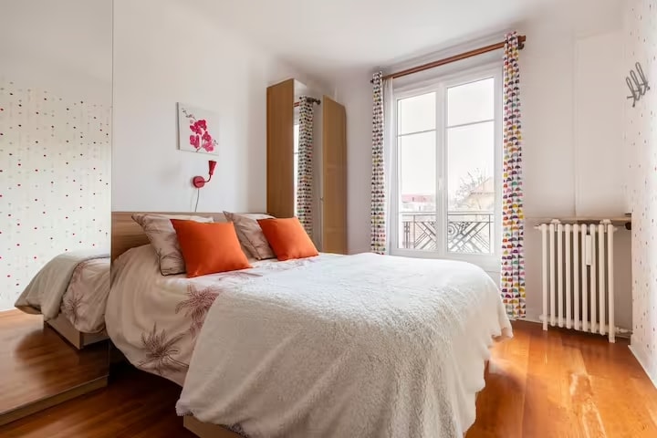 The bedroom features a large bed topped with decorative pillows and a cozy blanket. Natural light enters through double-glazed windows, reflecting off a mirrored wardrobe. A colorful curtain adds a pop of color, while a small piece of wall art complements the bright ambiance.