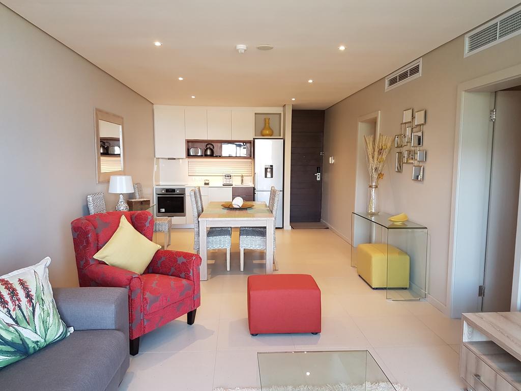 The living and dining area features a modern kitchen equipped with stainless steel appliances. A red armchair and yellow cushions complement the neutral-toned furnishings. A glass coffee table and dining table add functionality to the open space, with natural light illuminating the room.
