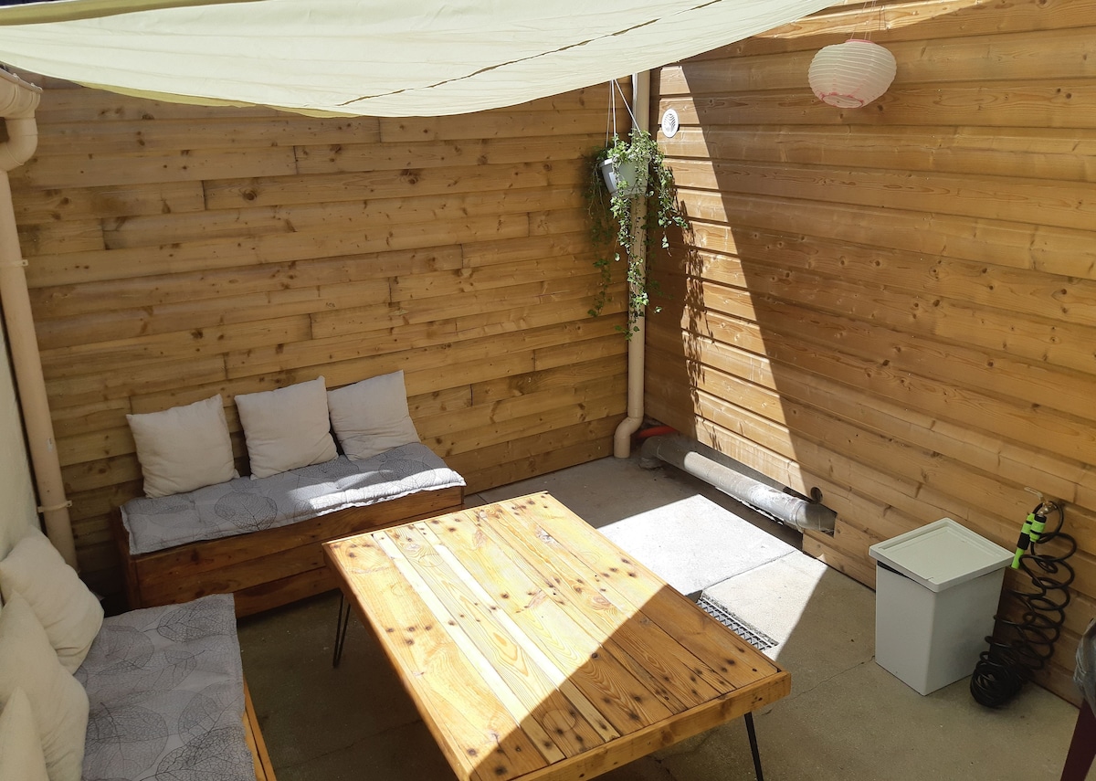 A private patio area features a wooden table surrounded by seating cushions against a backdrop of wooden walls. Natural light filters in, creating a relaxing setting. A green plant hangs from the wall, and practical items like a storage box and a hose are also visible.