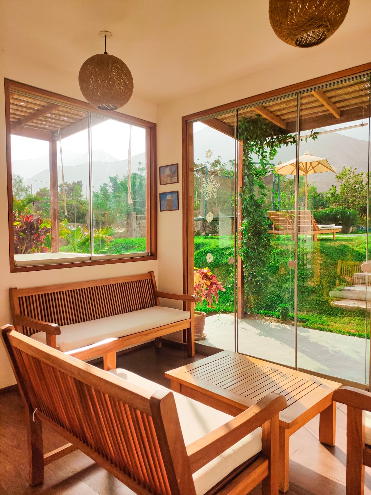 A sunlit corner of the house features a seating area with wooden furniture, including a table and two benches. Large glass windows allow for abundant natural light, offering views of the garden and outdoor seating, enhancing the connection with nature.
