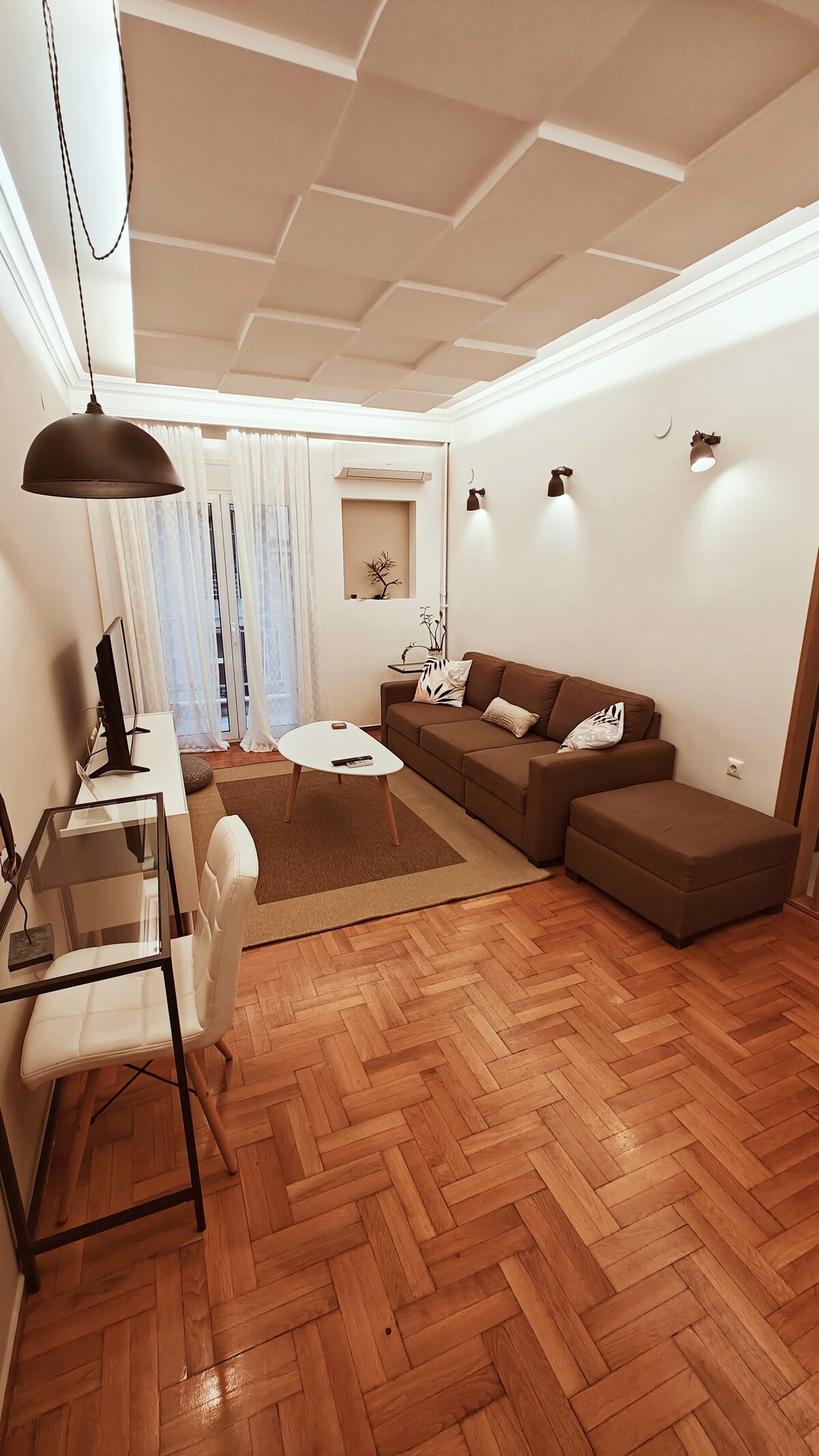 A modern living area features warm wooden flooring and a neutral colour palette. A comfortable sofa with pillows faces a TV mounted on the wall. A small table sits centrally, and natural light filters through sheer curtains, illuminating the space.