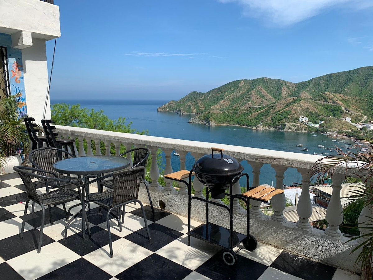 A spacious terrace is presented with a black and white checkered floor, featuring a round table and four chairs. A portable grill is positioned nearby, with stunning views of the turquoise bay and surrounding mountains visible in the background.