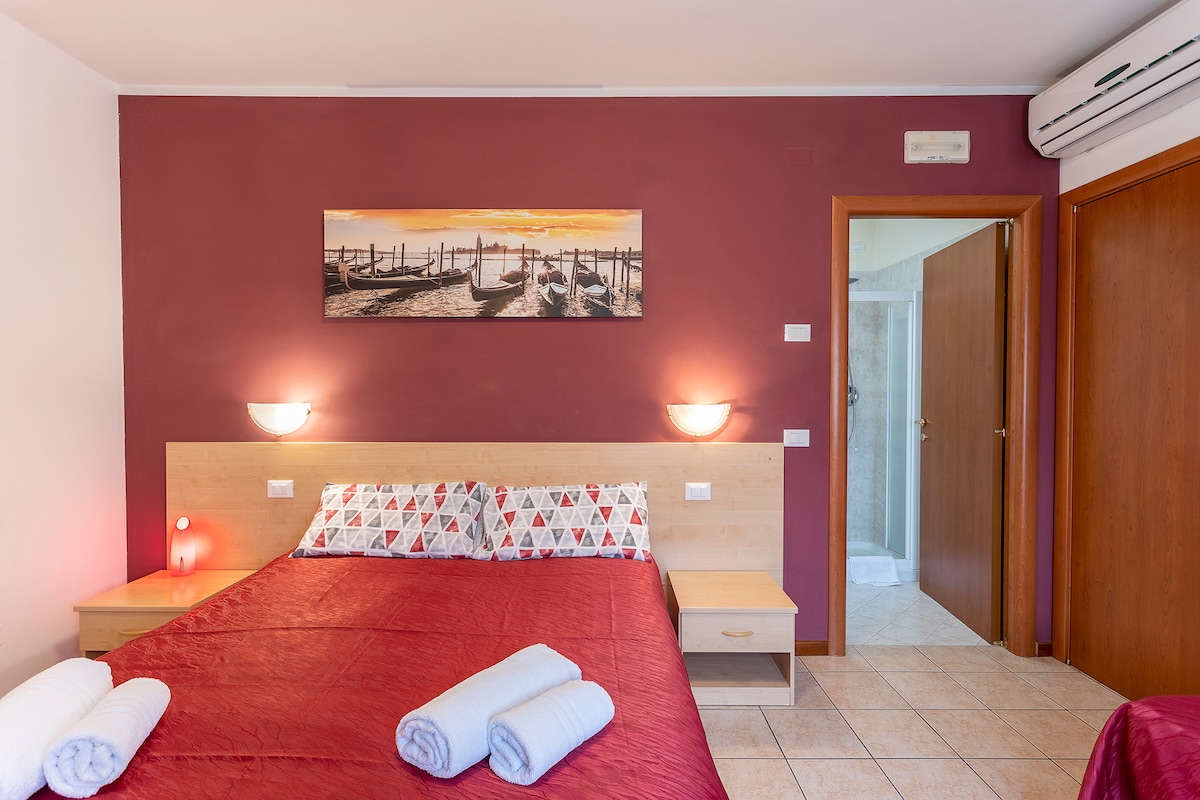 A comfortable bedroom showcases a spacious double bed adorned with a red quilt and patterned pillows. Soft lighting is provided by wall sconces, and a vibrant photograph of a sunset over water adds interest to the wall. A door leads to the bathroom in the background.