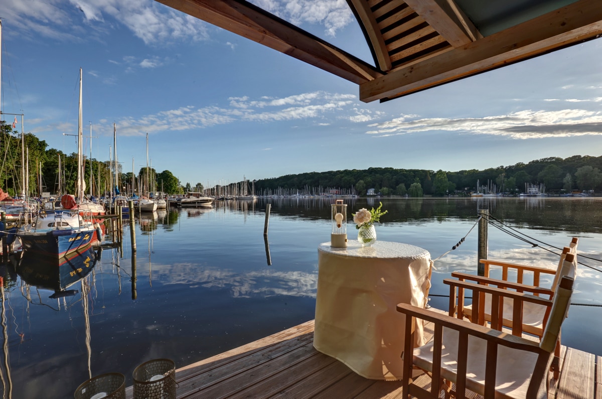 A serene view of calm waters reflecting a clear blue sky, with sailboats anchored in the distance. A small, elegantly set table is positioned beside a wooden chair, inviting relaxation and enjoyment of the tranquil surroundings.