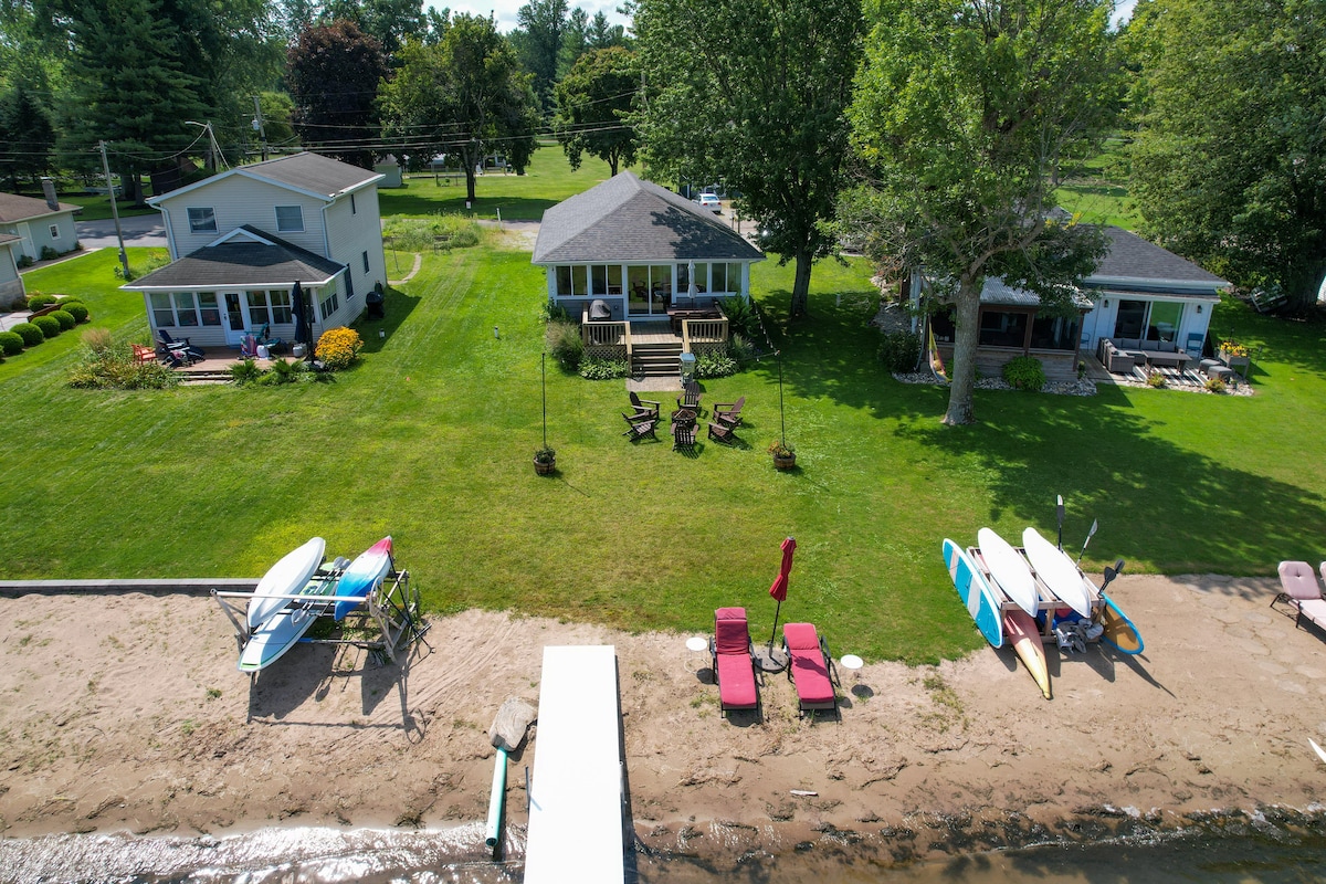 An aerial view captures three cottages set along a sandy lakefront, surrounded by green lawns and mature trees. Kayaks and paddle boards are neatly arranged on the beach, while seating areas are visible nearby for outdoor relaxation.