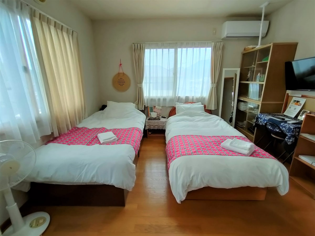 The guest room features two comfortable single beds, each dressed with fresh bedding and towels. Natural light pours in through large windows covered by sheer curtains, illuminating the warm wooden floor. A small seating area and a television are also present, enhancing the room's functionality.