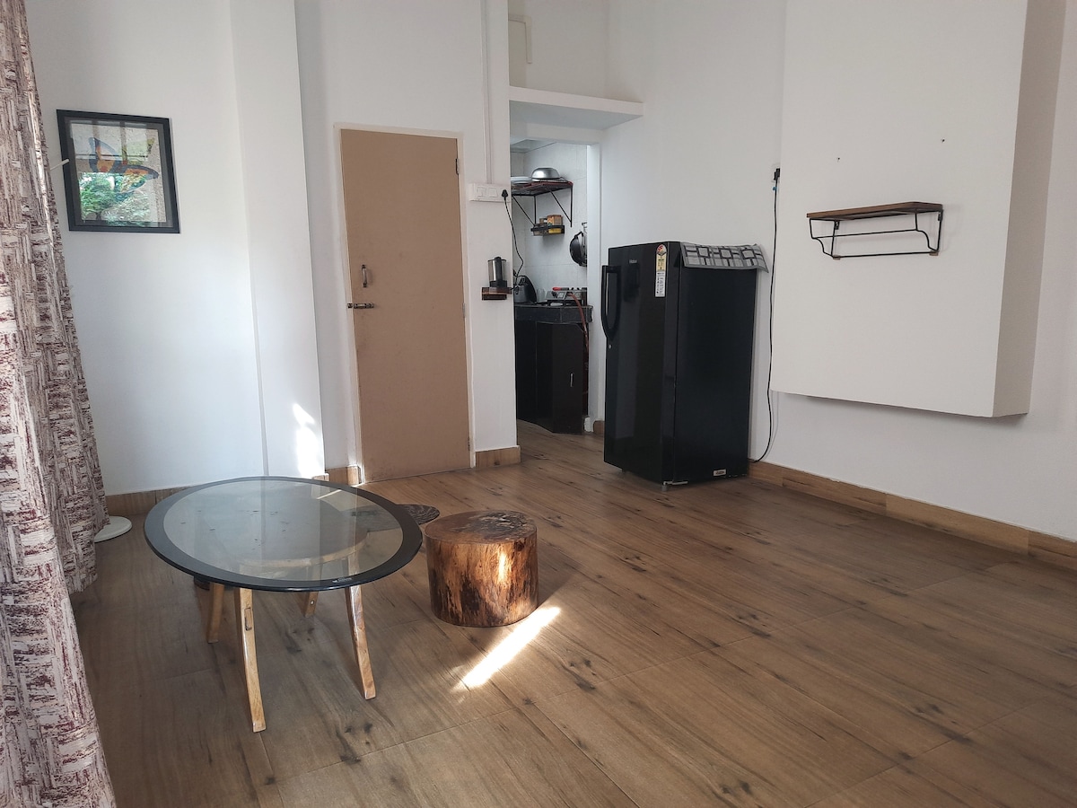 A spacious room features wooden flooring with a round glass table and a stump serving as a natural accent. A black refrigerator is visible in a kitchenette area, while natural light enhances the simple decor. Curtains frame a window, adding a touch of privacy.