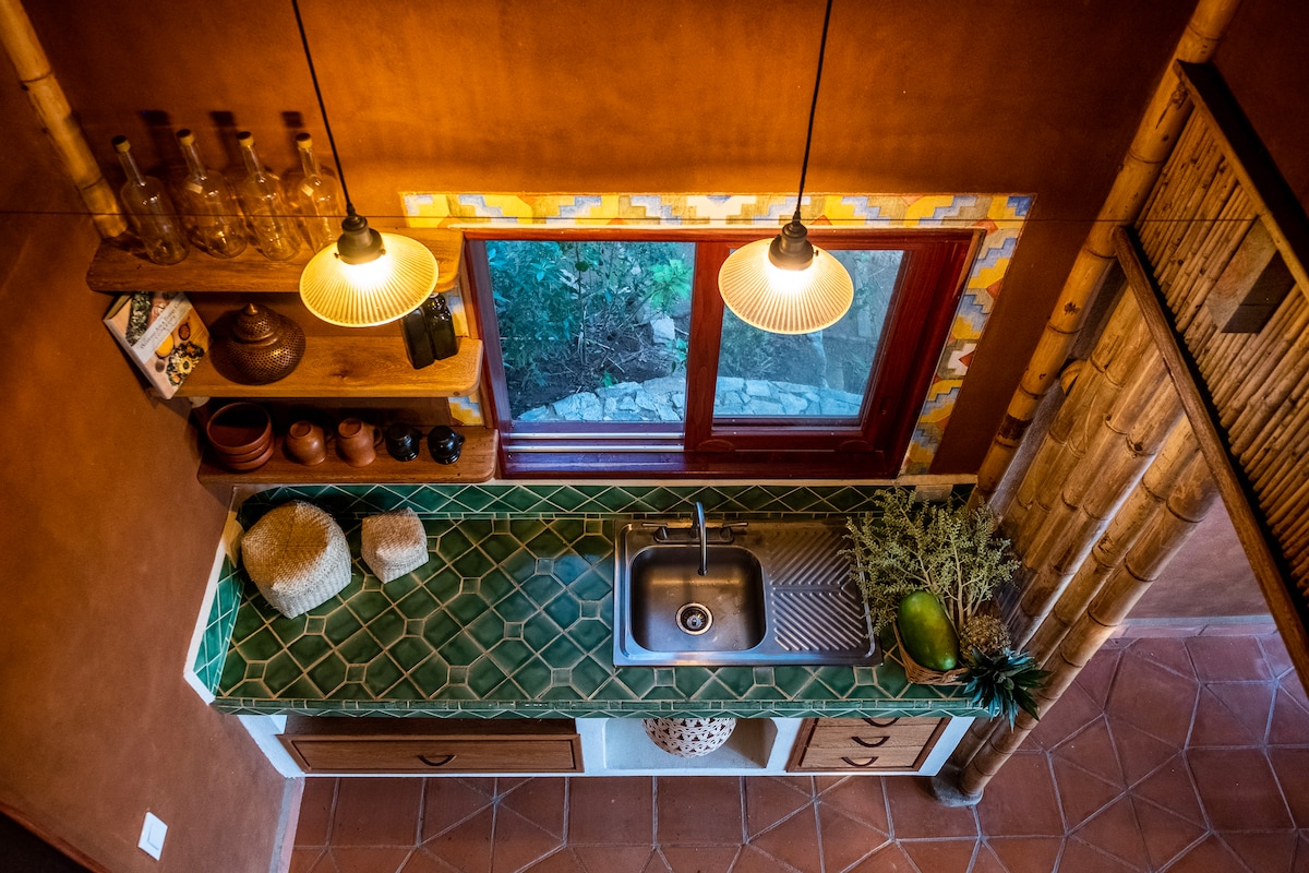 An overhead view of a rustic kitchen showcases a green-tiled countertop with a stainless steel sink. Shelves above display various jars and handcrafted items. Warm lighting is provided by two pendant lights, illuminating the space and a nearby window that offers a glimpse of the surrounding greenery.